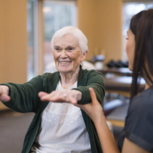 A female occupational therapist of Asian descent works with a cheerful senior female as she does exercises on an exercise ball. She is helping steady her as she holds her arms out.
